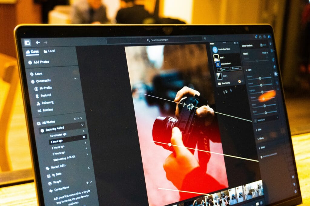 Laptop screen showing photo editing software with masking tools and light adjustment sliders open on an image of hands holding a camera